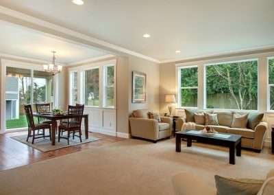 A well-lit dining area with large windows, a wooden dining table set, and beige carpet extending from the adjacent living room.