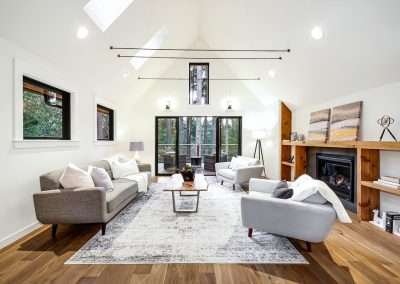 A bright living room with large windows, modern furniture, and a fireplace, opening to a deck with forest views.