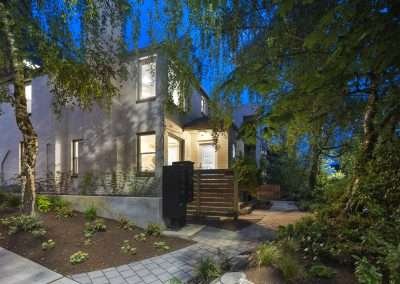 The exterior of a house at dusk, surrounded by greenery and a well-lit pathway.
