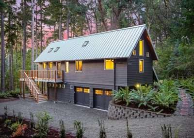 The front of a modern cabin with a gravel driveway, wooden deck, and metal roof, surrounded by forest trees.