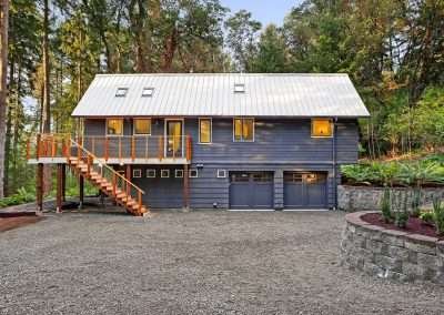 The exterior of a modern cabin with a white metal roof and wooden deck, surrounded by tall forest trees and lush greenery.