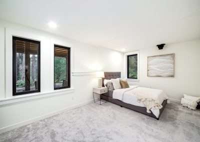 A bright guest bedroom with a plush bed, bedside lamp, and windows overlooking the forest, featuring soft carpeting.