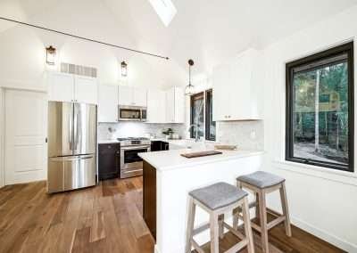 A modern kitchen with stainless steel appliances, dark wood cabinets, and white countertops, featuring large windows with forest views.
