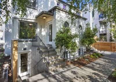 The main entrance of the home featuring a white brick exterior, a stairway, and surrounding greenery