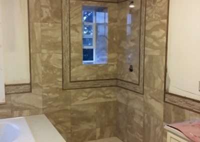 Bathroom renovation featuring beige tile walls and floors, a corner shower with mosaic accents, and a freestanding bathtub.
