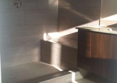 Bathroom renovation in progress with gray tile walls and floor, a large window, raised bathtub platform, and dark wood vanity cabinets.