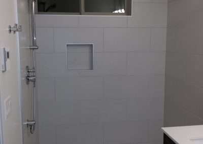A spacious walk-in shower with large white tiles, a single showerhead, and a built-in shelf.